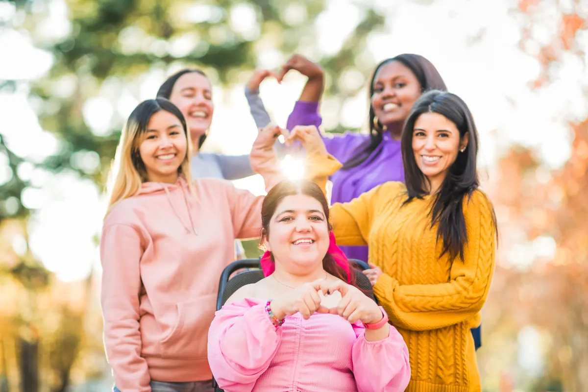A diverse group of smiling women in a sunlit park, including a young woman using a wheelchair, making heart shapes with their hands.