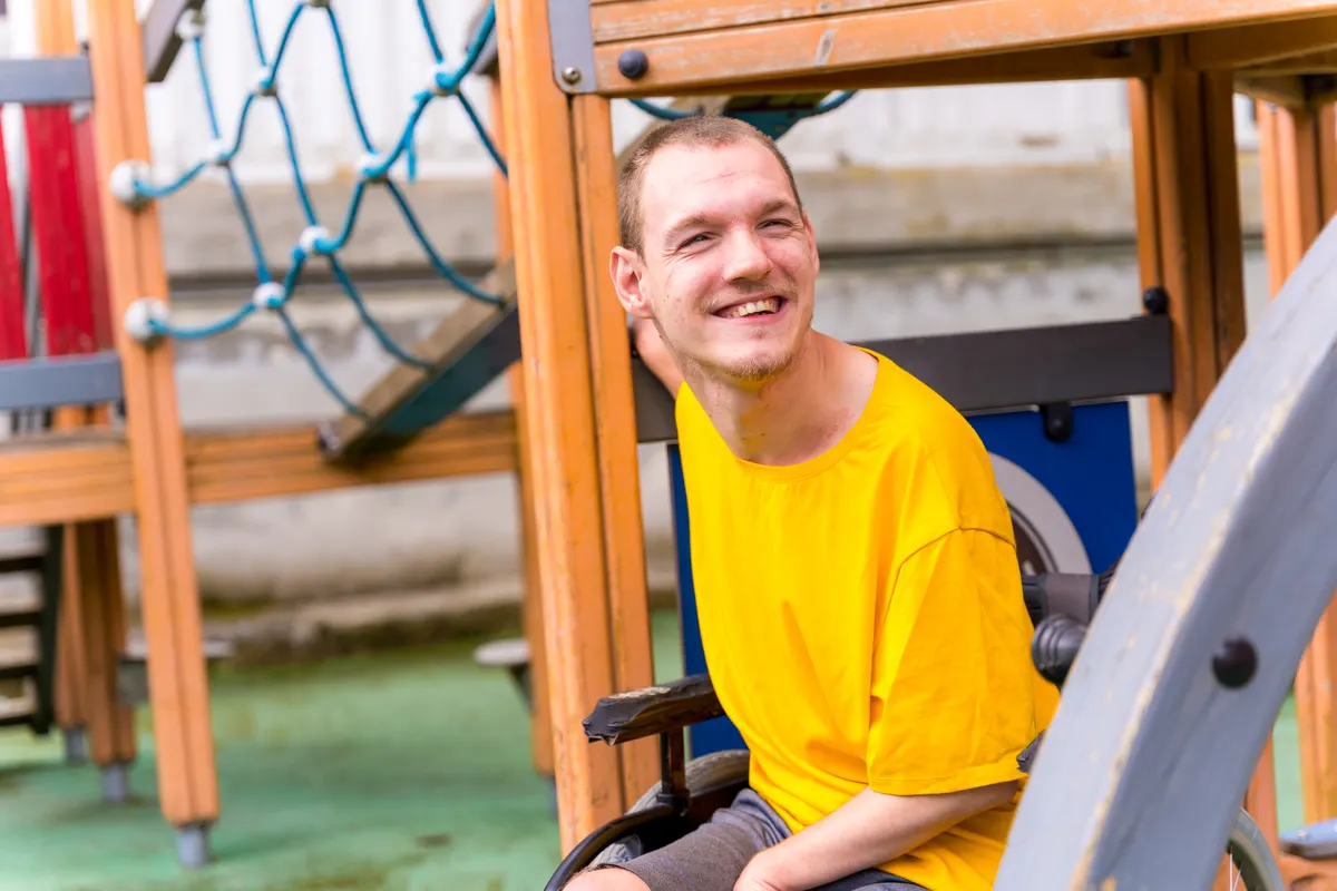 A young man using a wheelchair smiling warmly at an outdoor play space.
