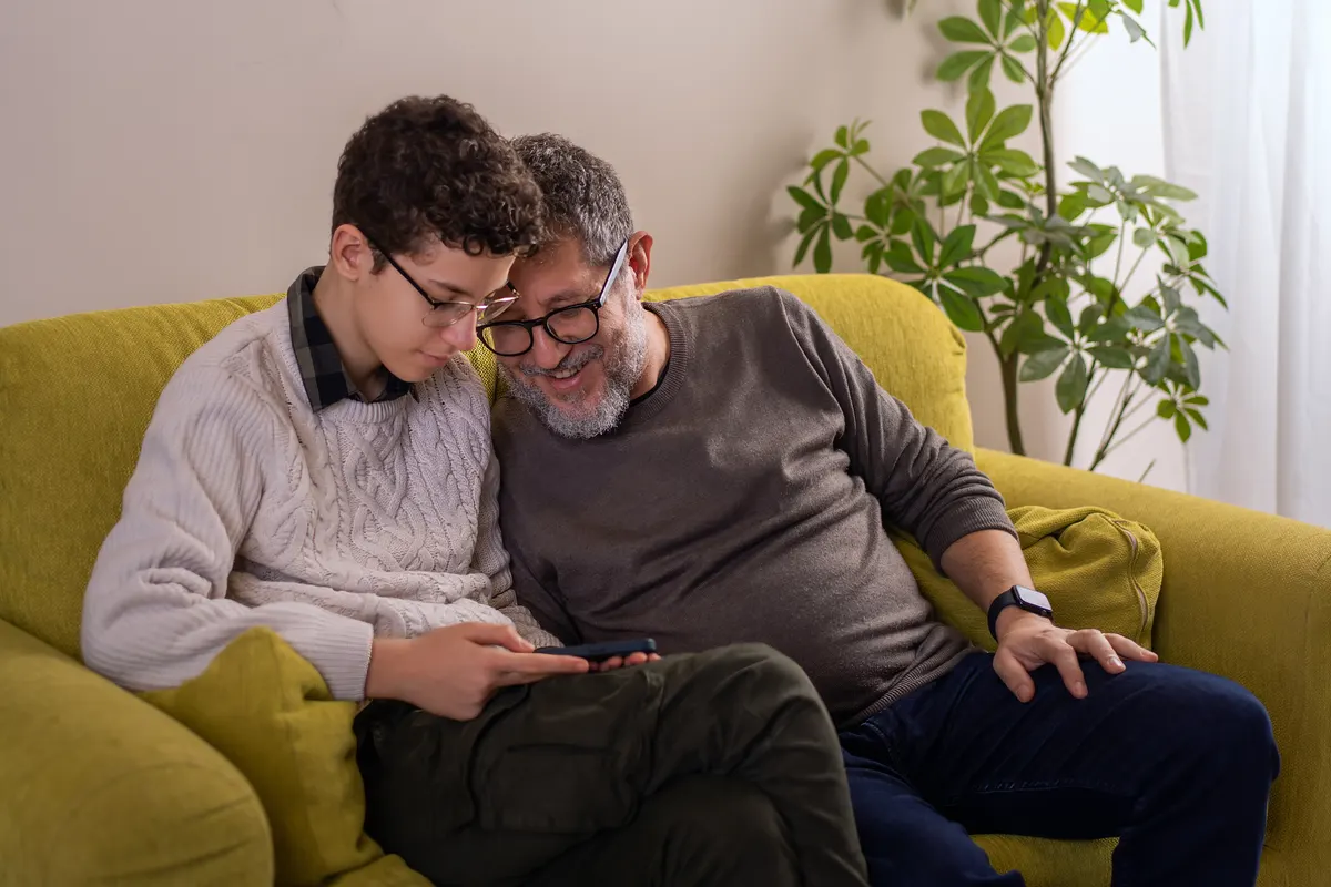An older man and a teenager sitting close together on a sofa, looking at a phone in a relaxed home setting.